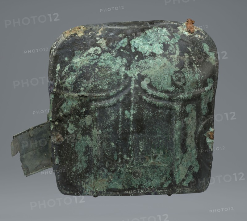 Two-piece torso armor with buckles, 4th century BC 375–325 BC, Southern Italy, weaponry; maker, two-piece cuirass design., An aged metal object with a rectangular rounded shape sits against a neutral gray background, showing a patina of green and teal corrosion mottled over dark gray and almost black areas; the surface has irregular patches of lighter green, turquoise, and pale mint along with small rusty brown spots and flaking where the metal has worn away, including a rust-colored crust near the top edge and a torn, bent tab protruding from one side that shows similar green and gray tones; the front displays subtle raised curved ridges and shallow indentations, and the edges are worn and uneven with small holes and rough textures.