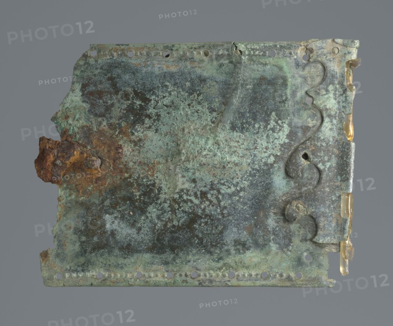 Two-piece torso armor with buckles, 4th century BC 375–325 BC, Southern Italy, weaponry, A rectangular corroded metal plate with irregular edges and a few small holes along two sides, showing mottled green and turquoise patina mixed with dark gray and brown rusted areas; a rough rusty brown protrusion is visible on the left edge, and a scalloped decorative metal strip with similar greenish corrosion runs near the right edge, where small translucent yellowish fragments cling to the edge; the background is a uniform medium gray.