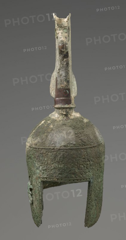 Bronze helmet, 8th–7th century BC, Italy, armament; height 46 cm, width 17.5 cm, depth 23 cm, weight 2.740 kg, A weathered metallic object with a rounded dome and elongated cheek guards, showing a rough, pitted surface texture and areas of corrosion; the top features a vertical crest or ornament resembling an animal head with a pronounced snout and flared ears, attached to a short neck above the dome; the metal shows a range of colors including greenish patina, brownish and dark brown patches, and some lighter gray and off-white encrustations; the open lower edge of the helmet has a rectangular face opening and small perforations along the rim, and the whole piece rests against a plain neutral gray background.