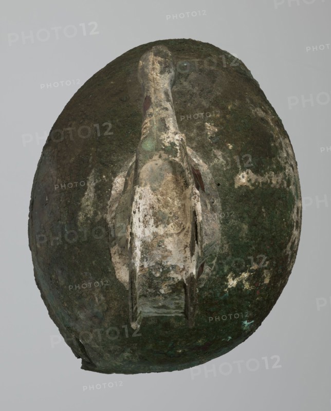 Bronze helmet, 8th/7th century BC, Italy, armament; maker, height 46 cm, width 17.5 cm, depth 23 cm, weight 2.740 kg, An oval object with a weathered surface showing a mix of dark green, brown, beige, and pale off-white areas, featuring a central raised ridge or handle-like protrusion that is lighter in color with patches of green and beige, and several irregular rough textures and mottled discolored spots across the surface.
