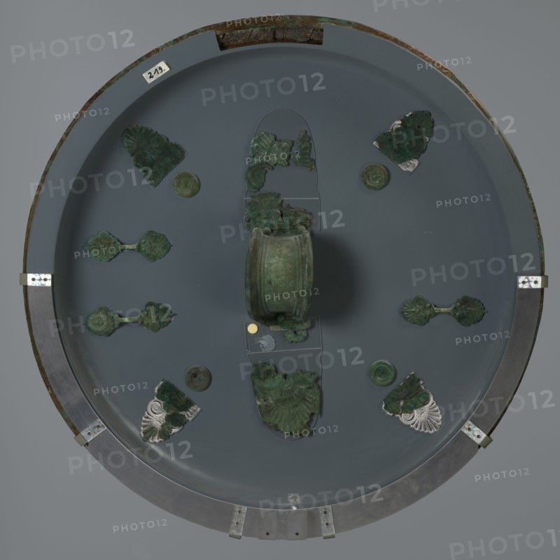 Hoplite shield, 6th century BC, Italy, weaponry, A round display with a muted gray background holds a group of greenish metal fragments arranged symmetrically around a central cylindrical object; the metal pieces show varied green patina and some darker verdigris, with a few fragments that include areas of silvery metal. The central cylindrical piece is green with mottled darker spots. Around it are several decorative pieces with scalloped, floral, and fan-like shapes in green tones, some attached to small round green discs. The display rim is metallic with a darker outer edge and small metal brackets, showing brownish corrosion in places; the overall colors visible are shades of gray, green, dark green, silver, brown, and a small circular spot of gold-toned color.