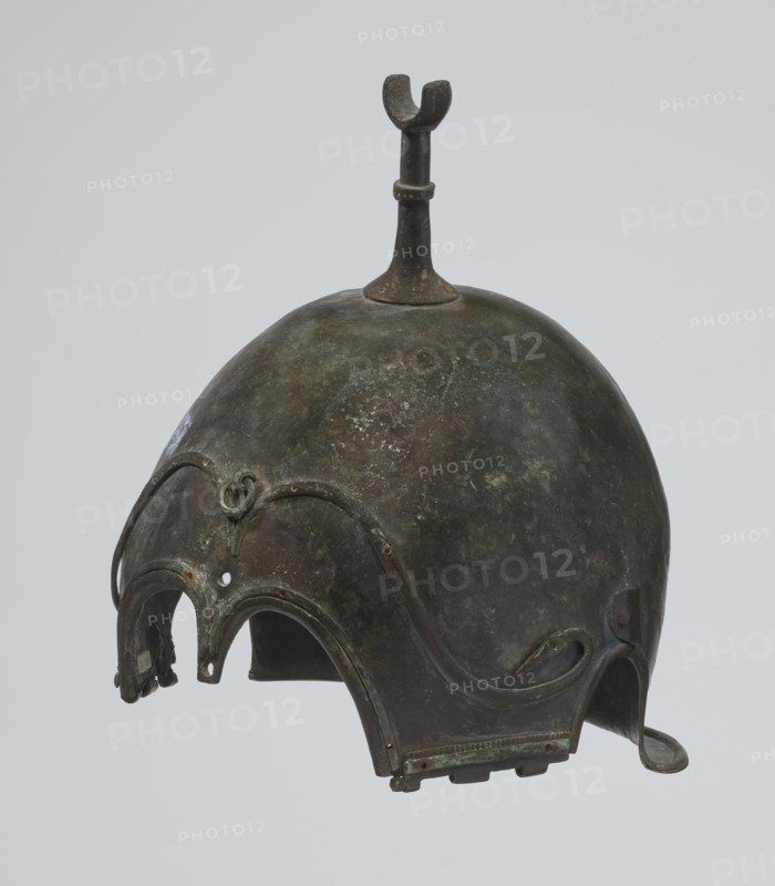 Bronze helmet, 4th–3rd century BC, Italy, armament; height 26.5 cm without cheek guards, width 19.8 cm, depth 21.6 cm, weight 0.758 kg without cheek guards., A rounded metal helmet with a domed top and a vertical fin-like projection rising from its crown, the surface showing mottled dark green, brown, and black patina with areas of lighter green corrosion; the front edge has an ornate scalloped rim with a few arching cutouts and curved decorative ridges, including a small raised loop detail near the forehead; the lower edges feature attached flaps and tabs with worn greenish and brown tones and a small hooked extension on one side; the overall texture appears aged and uneven with patches of darker and lighter coloration against a plain light gray background.