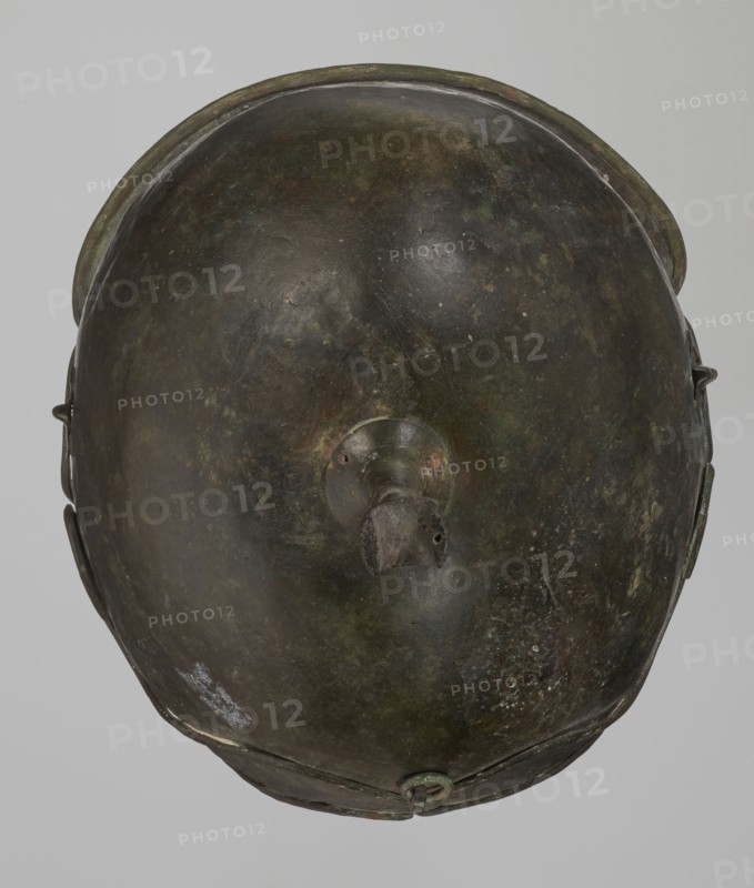 Bronze helmet, 4th–3rd century BC, Italy, armament; height 26.5 cm without cheek guards, width 19.8 cm, depth 21.6 cm, weight 0.758 kg without cheek guards., A top-down view of a rounded metal object with a smooth domed surface showing a patina of dark brown, green, and hints of black, with areas of lighter greenish corrosion and small pale speckles; a raised circular boss sits near the center with a short protruding stub that appears darker and rougher; the rim around the object is slightly raised and shows similar brown and green tones, and a few small attachments or fittings are visible along the sides and lower edge in matching dark metal colors.