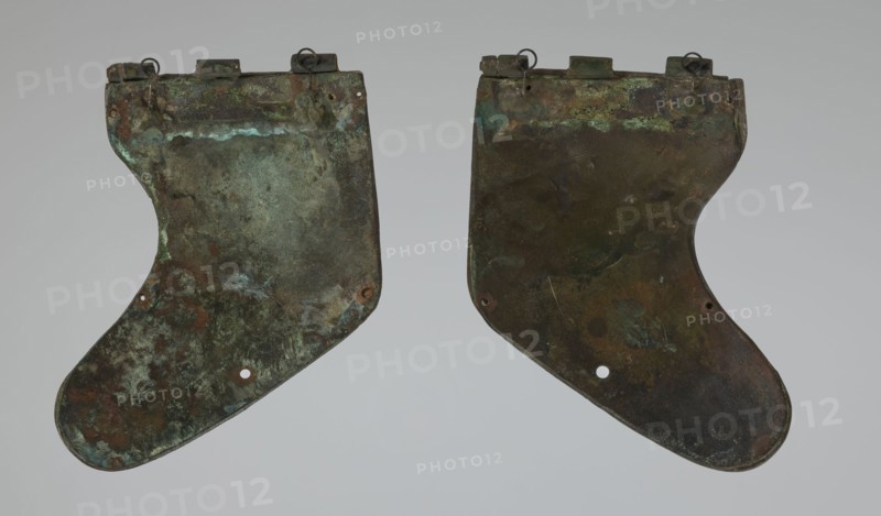 Bronze helmet, 4th–3rd century BC, Italy, armament; maker, height 26.5 cm without cheek guards, width 19.8 cm, depth 21.6 cm, weight 0.758 kg without cheek guards., A pair of irregularly shaped flat metal plates with curved lower edges and notched tops, shown side by side against a light gray background; the plates have a weathered, mottled surface with predominant greenish patina, dark brown and rusty orange areas, and touches of bluish-green and black; each plate has small circular holes near the lower portion and small metal loops or rings attached along the top edge; the surfaces show uneven textures, patches of corrosion, and variations in color intensity.