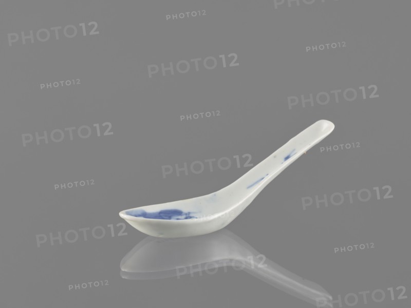 Spoon, bats iconography, ceramics, Harbin China, figurative scenes, blue and white porcelain, Qing style, Chinese art/culture, cutlery, Qing 1644–1911, 18th century, China, length 12.5 cm, A ceramic spoon with a curved handle rests on a reflective surface, showing a mirror-like reflection beneath it; the spoon is primarily white with a few blue brushlike marks on the bowl and along the handle, and the background and surface are shades of gray.