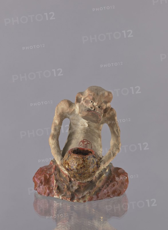 Figurine of a rakan Buddhist saint, ceramist, maker name: Choynowski, Józef 1833–ca.1915 author, Dawidow, Aleksander Pietrowicz 1838–1885 revindication, Japanese art; Japanese culture; Meiji 1868–1912; ceramics; figurines; male figurines; bowls; Buddhism; arhat iconography; rakan iconography; beggars; 1882 Meiji 15 ; Japan; sculpture; height 21.5 cm; width 19.0 cm, A small sculpted figure with a rough, textured surface in shades of beige, brown, and tan sits on a glossy reddish-brown base; the figure has an exaggerated facial expression with deep-set features and is holding a rounded, mottled bowl that shows dark brown and gold tones, while the background is a uniform light grayish-lavender color.