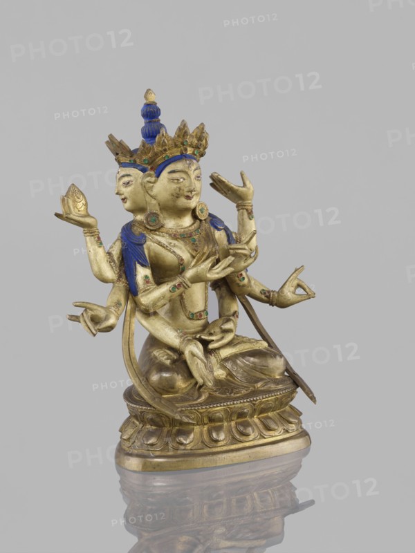 Ushnishavijaya, unknown author, 19th century, South China, or Tibet, bronze sculpture, statuette 17.0 cm high x 13.5 cm wide; lotus throne, mudras; Buddhist art iconography: Ushnishavijaya, Buddha, ushnisha. An identical figure is in New York., A gilt metal sculpture of a seated multi-armed figure with a secondary face at the back, showing a polished gold surface with blue painted hair and crown details and small green and red inlaid accents; the figure wears ornate jewelry and a tiered crown, holds various hand gestures and small objects, and sits on a lotus-style pedestal with carved petal motifs, all reflected faintly on a glossy surface beneath; visible colors include gold, blue, green, red, and a neutral gray background.