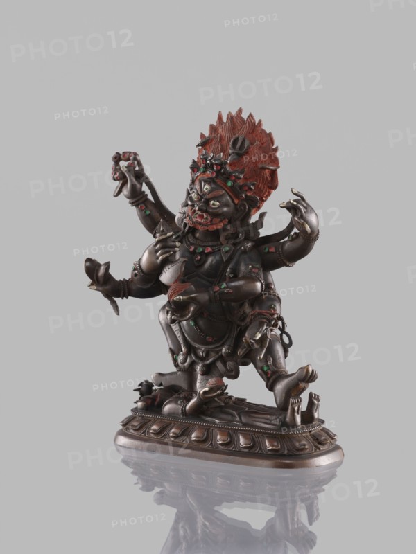 Six-armed Mahakala, dharmapala iconography, lotus throne, Tibetan Buddhist art, sculpture, 19th century, Tibet, height 16.4 cm., A dark bronze multi-armed figure mounted on an ornate base, with a textured red headdress and red highlights on the face and body; the figure has green and red decorative insets, multiple arms in varied gestures, and an expressive face with prominent eyes and teeth; the base is brownish bronze with carved ridges and the figure casts a faint reflection on a pale gray surface and background.