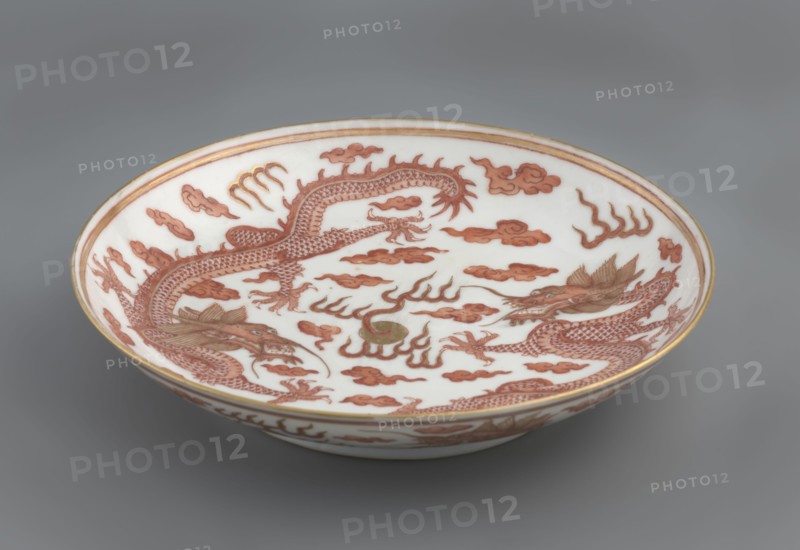 Plate, Chinese, Qing dynasty Guangxu, 1875–1908. Porcelain dish, height 3.2 cm, diameter 16.7 cm. Decorated in the mirror and on the outer wall with a pair of dragons with a pearl among clouds on a white background. Signed., A shallow round ceramic bowl sits against a gray background, showing an interior decorated with a pair of long, sinuous dragons rendered in reddish-brown tones with detailed scales, claws, and expressive open mouths, surrounded by stylized cloudlike shapes in the same reddish-brown; there is a small round element near the center with a muted greenish tone, and thin gold-colored bands rim the edge of the bowl while the bowl interior and exterior background remain white.
