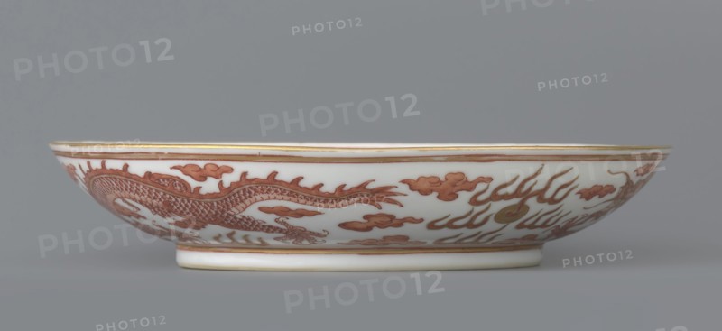 Plate, Porcelain dish, decorated in the mirror and on the outer wall with a pair of dragons with a pearl among clouds on a white background. Signed. Qing dynasty, Guangxu period 1875–1908. China. Height 3.2 cm; diameter 16.7 cm. Iconography: dragon, pearls, clouds., A shallow oval porcelain dish with a white base color and a band of reddish-brown decorative painting around its exterior showing a sinuous dragon motif and stylized cloud and flame shapes, accented with thin gold edging along the rim and base; the dish rests on a short foot and is photographed against a plain gray background.