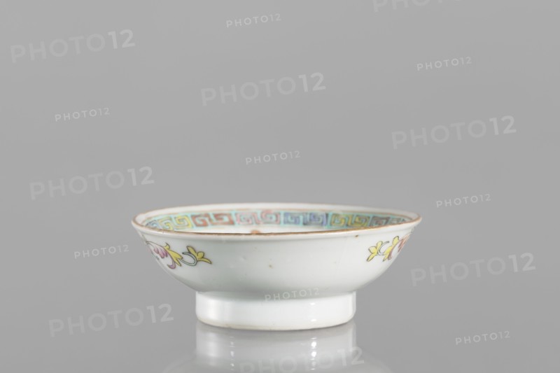 Bowl, gift, repossession Kraków; Chinese, Qing dynasty Guangxu period, 1875–1908, 2nd half of the 19th century; colored porcelain; vessel saucer, floral decorations, meander ornament; height 2.7 cm, diameter 7.5 cm; associated with Boxer Rebellion 1899–1901., A small shallow porcelain bowl sits on a reflective surface against a plain gray background, showing a white exterior with delicate painted floral motifs in yellow and pink with green accents on its sides, a faint brownish rim, and an inner border decorated with a repeating geometric band in pastel shades including blue, turquoise, yellow, and pink.