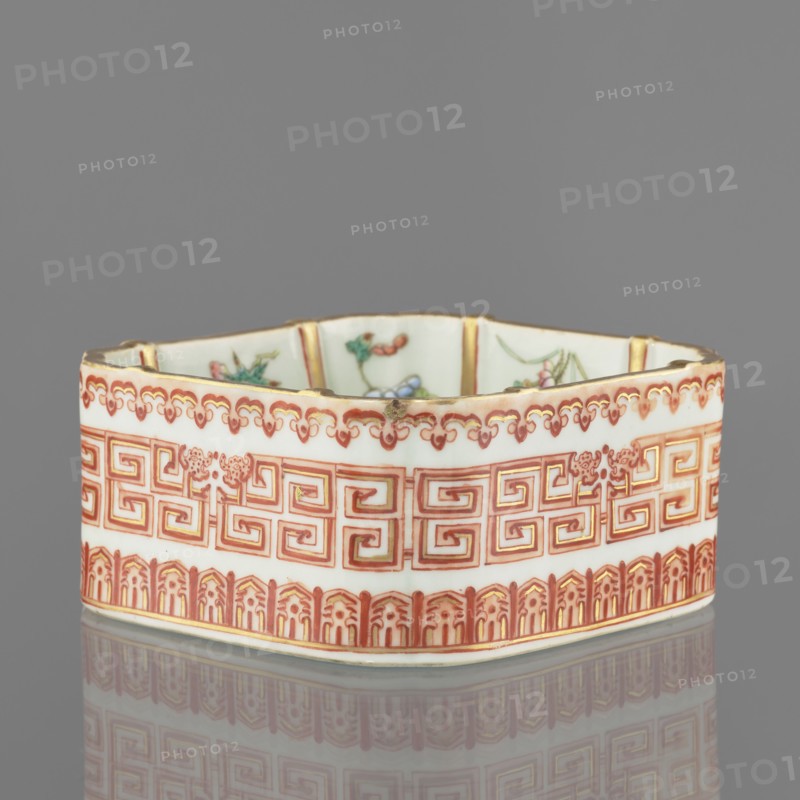 Vessel, Chinese, Qing dynasty Daoguang, 1821–1850. Ceramic colored porcelain vessel decorated on the outside with floral decorations, lambrequin pattern and meander strip. Height 6.0 cm; width 12.8 cm., A shallow hexagonal ceramic dish with an off-white base color decorated with intricate red and gold geometric and floral patterns around its exterior, a scalloped red and gold border near the top edge, and a band of red architectural-style motifs along the bottom; the interior shows hints of painted floral motifs in green, pink, and blue; the rim has small areas of wear revealing darker spots, and the dish sits on a reflective gray surface with a gray background.