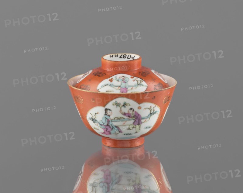 Bowl with a lid, ceramics, colored porcelain, Qing dynasty Tongzhi, 1862–1874, China; subject: bats and swastikas loosely placed in the background of the decoration, genre scenes; inscriptions: hieroglyphs; vessel; height 8.6 cm, diameter 10.3 cm; Chinese art., An orange ceramic bowl with a lid sits centered on a reflective gray surface, showing a reflection beneath it; the bowl has white cloud-shaped panels decorated with painted figures in pastel colors including light blue, pink, purple, green, and flesh tones, and small decorative motifs in gold on the orange background; the lid has a short orange neck and a white interior rim, and the overall scene is set against a plain gray background.