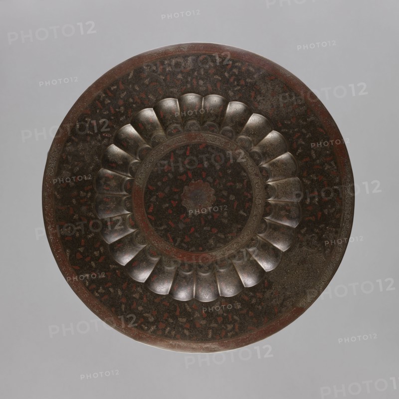 Tray, South Asian, Indian, brass tray with a separate facet; engraved and painted decoration depicting a central rosette and floral ornaments placed around it; floral and geometric ornamentation; 19th–20th century, India; vessel; height 2.0 cm, diameter 59.0 cm, A round decorative object viewed from above with a scalloped inner ring and concentric circular patterns, showing a dark, metallic surface with a mottled texture and visible patches of red and brown; the scalloped sections have a slightly reflective, silvery-grey appearance, while the surrounding areas include darker blackish-brown tones and hints of rusty red, all set against a plain light grey background.