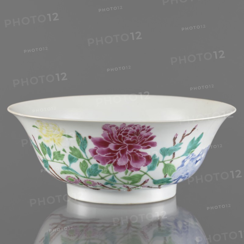 Bowl, porcelain, semicircular body widening toward the upper edge; white, transparent, fritted glaze with overglaze enamel-painted decoration of rocks and peonies in colored porcelain; Qing dynasty, Yongzheng period 1723–1735, China; height 7.6 cm, diameter 19.5 cm, weight 0.3 kg., A shallow white porcelain bowl with a flared rim sits against a gray background, decorated around its exterior with painted floral motifs including a large deep magenta flower, a pale yellow blossom, and a smaller blue flower, connected by green leaves and brown stems; the bowl rests on a short foot and casts a subtle reflection on the surface below, and the visible colors include white, gray, magenta, yellow, blue, green, and brown.