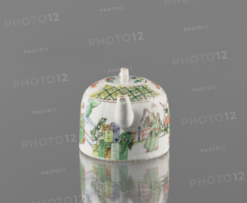 Teapot with lid, ceramics, figurative scenes, Paderewski, Ignacy Jan 1860-1941, gardens, Chinese art, Chinese culture, colored porcelain teapot, porcelain fragment; white, transparent glaze. Decoration painted with overglaze enamel paints and iron red. 19th century, China. Vessel; height 8 cm, width 14.3 cm, diameter 9.3 cm., A small rounded ceramic teapot with a short spout and a lid sits on a reflective surface, showing a mirrored reflection beneath. The teapot is predominantly white with painted scenes and decorative patterns in green, orange, yellow, blue, and black. One side shows a figure wearing a green robe, and other painted figures and architectural shapes appear around the body. The lid has a small white knob and a patterned band with green and yellow geometric details, and there are floral or decorative accents in orange and green on the top. The background is a uniform gray.