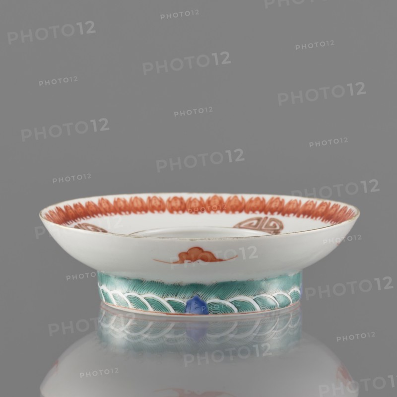 Bowl stand. Porcelain shard in the form of a ring with white glaze. Decoration painted with overglaze iron red, blue and green enamel, including bats and shou longevity hieroglyphs. Colored porcelain stand, Qing dynasty, 19th century, China. Height 3.3 cm; diameter 12.5 cm., Classification: Chinese ceramics., A shallow ceramic dish with a flared rim resting on a short foot, shown against a gray background. The upper surface of the dish is glossy white with a decorative band of reddish-orange petal-like shapes around the inner rim and a small reddish-orange cloud motif on the outer wall. The foot is painted in shades of green with a repeating wave pattern outlined in white, and there is a small blue accent where the foot meets the base. The edge of the rim has a thin warm-toned line, and the dish sits on a reflective surface that mirrors its colors. The visible colors include white, reddish-orange, green, blue, warm brown, and gray.