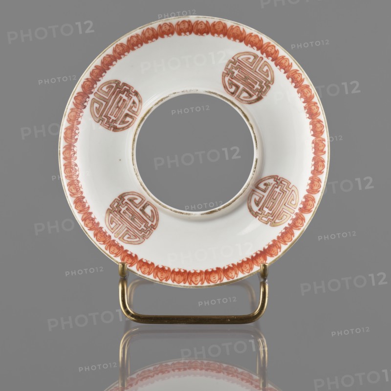 Bowl stand. Porcelain shard in the form of a ring with white glaze; decoration painted with overglaze iron red, blue, and green enamel. Decorated with bats and shou longevity hieroglyphs. Colored porcelain; Qing dynasty, 19th century, China. Height 3.3 cm; diameter 12.5 cm., A white round ceramic ring with a central circular opening sits upright on a small metallic stand; the ceramic has a decorative border in a reddish-orange hue composed of repeating floral or petal-like motifs and several circular medallion designs in the same reddish-orange color spaced around the inner area, with thin metallic or gold-colored trim along the inner and outer edges; the stand is brass or gold-toned and its reflection is visible on the smooth gray surface beneath, and the background is a uniform gray.