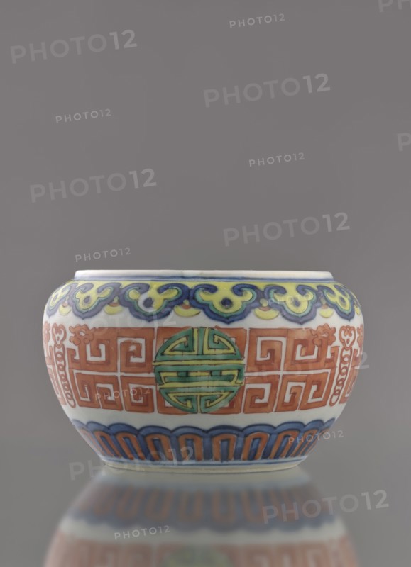 Globular vessel, porcelain body with white glaze; decoration painted underglaze with cobalt and overglaze with enamel paints and iron red. Low, flattened form without cover. Colored porcelain, Chinese, Qing dynasty 19th century. Decoration includes longevity shou iconography and meander ornament. Height 7.7 cm; diameter 10.3 cm; weight 0.35 kg., A rounded ceramic bowl with a glossy finish sits against a neutral gray background, showing a palette of white, blue, green, yellow, orange, and red; the rim area features a wavelike decorative band in blue and yellow, below which a central band displays repeating geometric motifs in orange interspersed with circular medallions in green; the lower section has an arched scallop pattern in blue and orange above a pale base, and a subtle reflection of the bowl appears on the surface beneath it.