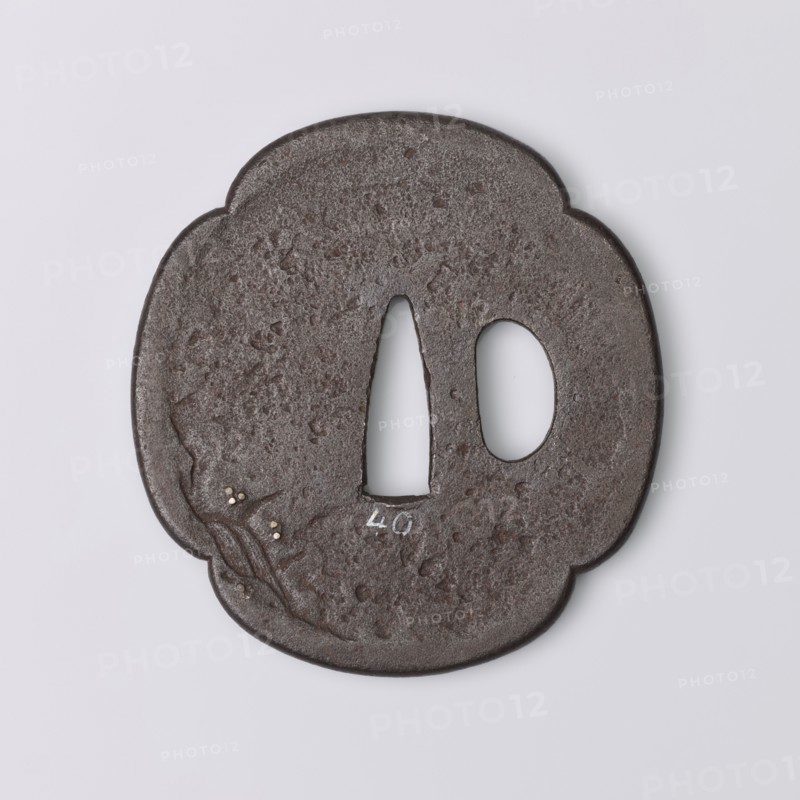 Crossguard tsuba, Kaneie,  school craftsman author. Japanese, Edo period 1603–1868, 18th century. Tsuba in the shape of a quatrefoil mokkô with an uchikaeshi edge, katahitsu ana one side hole. Decorated in relief showing the sennin Gama sitting under a tree, holding a staff and looking at a three-legged frog; on the reverse a sketched landscape motif with a waterfall. Details eyes, pebbles, staff inlaid with gold and shakudo. Origin: Fushimi, Yamashiro Province. Materials/subject: weapons — sword crossguard katana. Dimensions: height 7.2 cm, width 6.8 cm, depth 0.4 cm., A round, slightly irregular dark brown metal object with a textured, pitted surface and a scalloped outline, featuring two openings near the center—one elongated teardrop-shaped and one oval—and small raised decorative elements on one side with tiny pale inlays; the background is light gray and the visible colors include dark brown, pale off-white, and light gray.