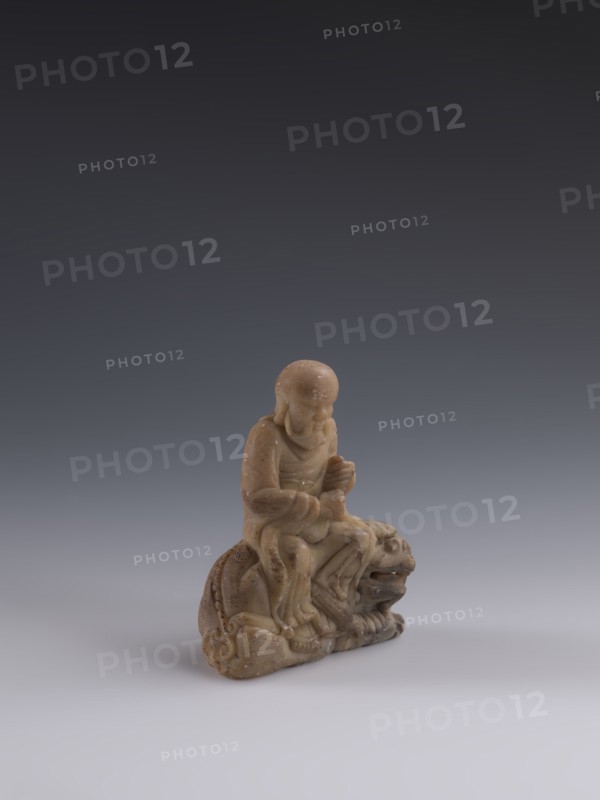 Luohan on a lion, arhat iconography, lion iconography, figurine, Chinese art, Chinese culture, Qing dynasty 1644–1911, Chinese Buddhism, 19th century, China, sculpture; height 9.3 cm, width 7.7 cm, depth 3.7 cm., A small carved figurine in shades of tan and light brown sits on a smooth pale gray surface against a gradient background that shifts from dark gray at the top to medium gray near the center; the figurine depicts a seated human figure with a rounded head and draped clothing, hands positioned in front, seated atop an animal-like form with a pronounced open mouth and rounded features; the carving shows smooth, worn textures and subtle speckled markings across its surface, with shadows beneath and to one side.