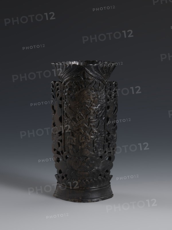 Vase, gift; Chinese, Qing dynasty 1644–1911; dark soapstone vase with bas-relief and openwork in places, decorated with dragons; one of three; 19th century; vessel; height 17.7 cm, width 8.7 cm., A tall, dark carved object with a flared top and a rounded base stands against a gradient gray background; the surface shows intricate openwork and relief carving with curving, floral and organic motifs, including petal-like shapes near the base and layered, leaf-like forms toward the rim, and the color palette is primarily dark brown to nearly black with subtle variations in sheen.