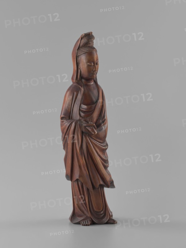 Guanyin Bodhisattva, unknown sculptor, gift, figurine, Chinese art, Chinese culture, goddess, Chinese Buddhism, Qing 1644–1911, bodhisattva iconography, wood carving, solid, polished, 19th century, China, sculpture, height 32 cm, A carved wooden figure of a robed person standing on a flat surface, depicted with closed eyes and a serene facial expression, wearing a hood or head covering with a carved detail at the top, draped garments with flowing folds and layered robes, hands held together at the waist, exposed feet peeking from beneath the robes; the wood shows warm brown tones with darker and lighter brown variations and subtle reddish-brown highlights against a plain neutral gray background.