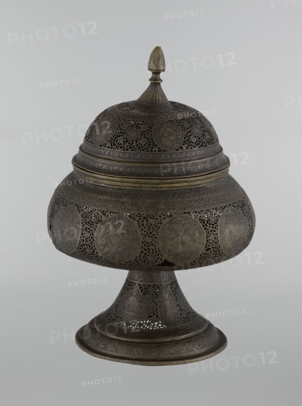 Censer, Persian Iran, Qajar dynasty late 18th–19th century. Brass censer, height 33.0 cm, diameter 22.0 cm. Decorated with figurative scenes and hunting iconography; human figures, animals, demons in cartouches; dense arabesque background; plant motifs; fantastic creatures and beasts dragons, fantastical beasts, riders, acrobatics; Islamic art, Persian decorative style., A domed, lidded metal object on a flared pedestal with elaborate openwork and engraved circular medallions, showing a dark brown patina with areas of darker and lighter brown and subtle greenish-brass highlights, featuring a pointed finial on top, layered bands and filigree patterns around the dome and pedestal, and intricate floral and geometric motifs throughout.
