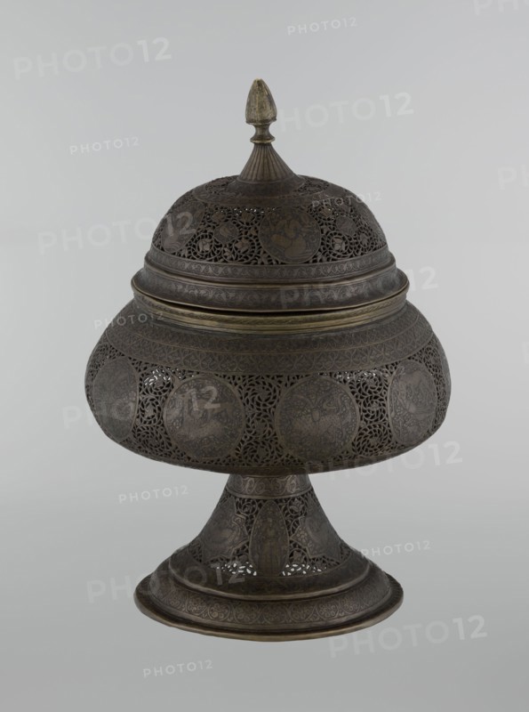 Censer, Persian, Qajar dynasty 1785–1925, 18th–19th century; Iran. Brass censer, height 33.0 cm, diameter 22.0 cm. Decoration: figurative scenes of hunting, human figures, animals, demons in cartouches; dense arabesque background with plant motifs, fantastic creatures dragons, beasts, riders, acrobatics; Islamic art., A decorative metal vessel with a lid and pedestal base sits against a plain light gray background; the vessel is dark brown with areas of muted brass tones and features intricate openwork filigree and rounded medallion panels around its bulbous midsection and dome-shaped lid, and the lid is topped by a small finial that shows a slightly lighter brass color, while the pedestal and rim display repeating engraved patterns and the overall surface shows a subtle aged patina.