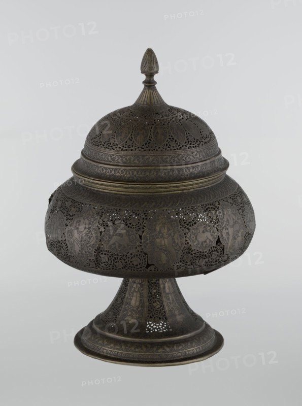 Censer, Persian, Qajar dynasty late 18th–19th century, Iran. Brass censer with figurative scenes of hunting, riders, acrobatics, dragons and fantastic beasts, human figures, animals and demons in cartouches, dense arabesque background and plant motifs; height 33.0 cm, diameter 22.0 cm., An ornate metal lidded vessel with a rounded body and pedestal base, displaying intricate openwork and pierced patterns; the lid has a pointed finial and layered decorative bands, and the body shows a series of framed figural and animal motifs within delicate filigree; the pedestal flares outward and repeats the pierced and engraved decoration; visible colors include dark brown, bronze, and hints of muted gold against a pale gray background.