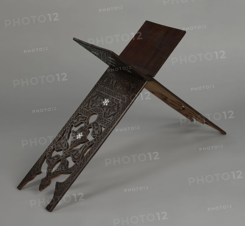 Quran lectern rahla, Ottoman Turkish, 19th century. Wooden piece of furniture used for holding the Quran, decorated with arabesques, geometric ornament, rosettes, lotus tulip motifs, lotuses, and tulips. Decorated with rosettes, geometric ornament, arabesque, lotus tulip motif. Unfolded: height 50.6 cm, width 91.5 cm, depth 21.3 cm, weight 1.7 kg. Folded: height 80.5 cm, width 21.3 cm, weight 1.7 kg., A carved wooden book stand formed by two interlocking panels arranged in an X shape, showing intricate openwork and relief carvings with floral and geometric motifs; the wood is dark brown with areas of lighter brown on the underside and edges, and there are small inlaid pieces that appear white; the background is a smooth gray surface and gray backdrop.