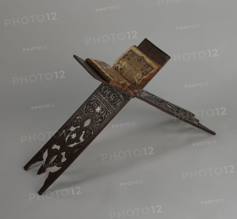 Quran lectern rahla, Ottoman Turkey, 19th century. Function: Quran lectern rahla; decorated with rosettes, geometric ornament, arabesques, lotus tulip motif;, Islamic, Turkish, Ottoman. Dimensions: unfolded: H 50.6 cm, W 91.5 cm, D 21.3 cm; folded: H 80.5 cm, W 21.3 cm; weight 1.7 kg., A carved wooden book stand with intricate openwork patterns holds an open illuminated manuscript; the stand is dark brown with visible carved floral and geometric designs, and small lighter inlay accents. The manuscript has aged beige to yellowed pages with ornate decorative borders in gold and blue and reddish tones, and a darker brown cover visible at the back. The background is a smooth neutral gray.