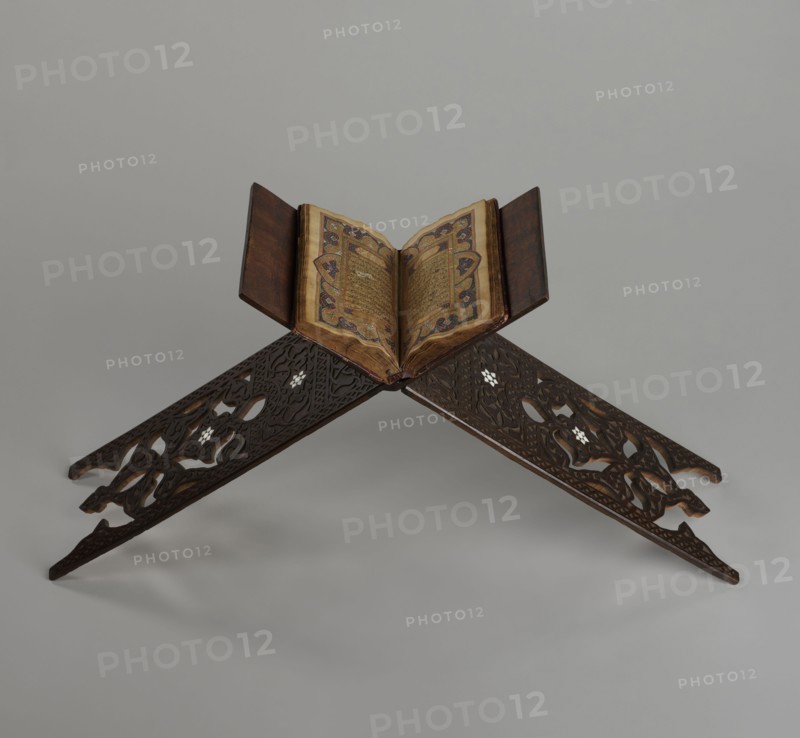 Quran lectern rahla, Islamic art, Turkish, Ottoman dynasty 1299–1922. Quran lectern decorated with arabesques, geometric ornament, rosettes, lotus tulip motif; decorated with rosettes. 19th century, Turkey. Piece of furniture. Dimensions unfolded: height 50.6 cm, width 91.5 cm, depth 21.3 cm, weight 1.7 kg; folded: height 80.5 cm, width 21.3 cm, weight 1.7 kg., An open book with ornate patterned page edges rests on a carved wooden book stand; the book's pages show intricate decoration in shades of gold, blue, red, and brown, and the covers are dark brown; the stand is dark wood with carved, interlacing floral and geometric cutouts and small inlaid lighter-colored accents, set against a neutral light gray background.