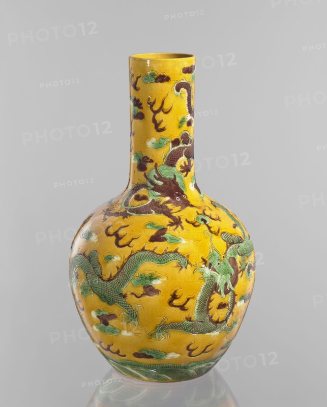 Vase, Chinese; Qing dynasty, Kangxi period 1662–1722; porcelain cylindrical-neck vessel with fa-hua decoration depicting dragons; height 41.0 cm, diameter 24.3 cm, weight 3.0 kg., A tall ceramic vase with a narrow neck and rounded body sits against a plain light gray background; the vase is predominantly yellow with decorative motifs in green, brown, and teal, featuring a group of sinuous dragon figures with scaled bodies and clawed limbs entwined around the surface, surrounded by stylized cloud-like shapes and flame-like elements, and a band of green wave patterns encircling the base; highlights and reflections on the glaze indicate a glossy finish.