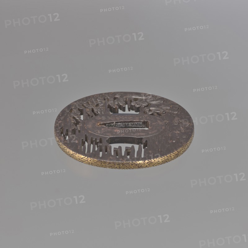 Crossguard tsuba, Takehara Tomomasa; oval tsuba tatemaru gata with rectangular edge, inlaid with gold using the nunome technique in a rinzu ornament. Side holes ryohitsu ana are part of the openwork background. Decorated with relief carving and engraving depicting two elderly people standing by the seashore beneath a spreading pine tree, gazing at the moon emerging from the fog and folding their hands in prayer; two brooms and several shells lie on the shoreline sand. The man is Jo, son of Izanagi; the woman is Uba — their legend Takasago symbolizes long life and a happy marriage. Japanese; Edo period, 18th century. Materials/subject: weapons — swords/katana crossguard tsuba; motifs: pine trees, water, moon, shells, sea waves, men's and woman's heads. Dimensions: height 7.0 cm, width 6.7 cm, depth 0.45 cm., A round metallic object with a dark brown surface and a decorative golden edge sits against a light gray background, showing carved raised and recessed patterns across its top inclu...