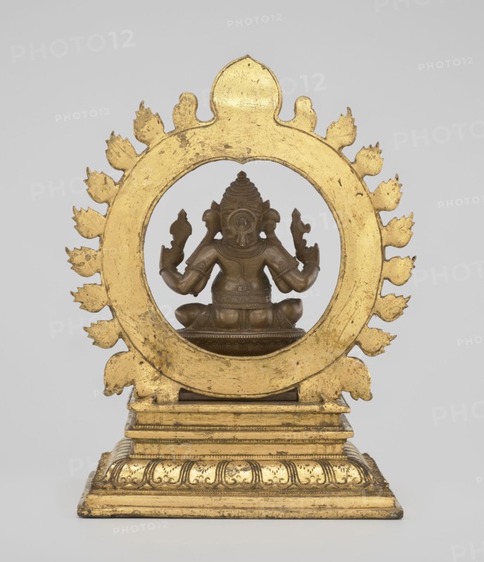 Ganesha, 19th century, India; sculpture; height 26.0 cm, width 17.5 cm. The deity sits on a double lotus throne placed on a rectangular flat base; the whole is mounted on a large, strongly profiled pedestal. At the back of the figure is a circle of fire. The figurine consists of three parts: a statuette inserted onto a pedestal and a fire ring inserted into the pedestal holes using two pins. Iconography: Indian/Hindu art — elephants, lotus throne, halos, birds, rats; attributes include crowns/tiaras, tusks, mounts, armor; associated with Hinduism and Shaivism., A gilded circular frame with a scalloped flame-like border surrounds a seated bronze figure viewed from the back; the frame and stepped base are gold with worn areas revealing darker tones, and the figure is a brownish bronze with detailed headgear, arm ornaments, and a seated posture on a small pedestal; the background is a pale gray.
