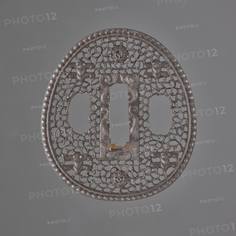 Crossguard tsuba, sword crossguard in namban style, openwork egg-shaped tsuba with a separated edge and pearling; rectangular seppa-dai decorated in low relief with a stylised volute ornament. Surface decorated with an openwork plant tendril karakusa with two sacred jewels tama woven into it and four mantis heads seen from the front. Not signed. 18th century Edo period, 1603–1868, Japan. Tsuba; height 6.5 cm, width 5.8 cm, depth 0.35 cm., A round, ornately pierced metal object with a raised beaded rim and an openwork lattice of interlocking curved shapes filling the interior; the central area features a vertically oriented rectangular opening framed by a carved border with symmetrical curved motifs and a small raised notch at its lower edge, and there are a few stylized animal-like figures integrated into the lattice around the central opening; the surface shows a metallic dark gray color with areas of slightly lighter gray highlights and a small patch of brownish-gold near the lower e...
