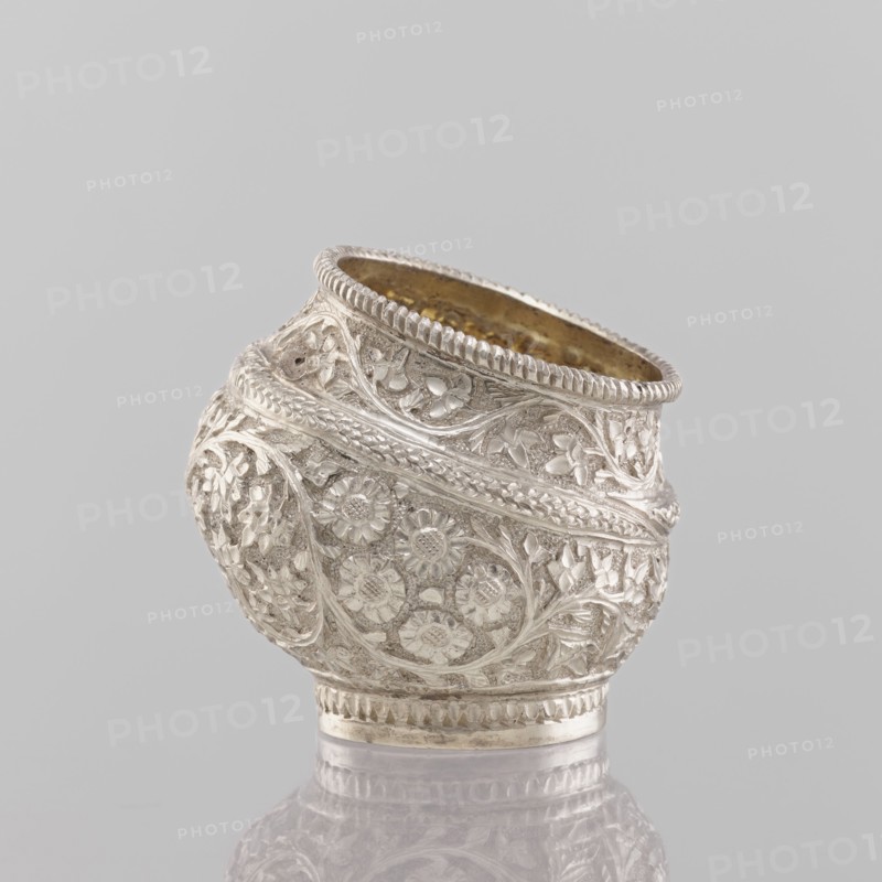 Bowl, Indian art, Indian culture, plant ornament, flowers, leaves, containers, applied art, cups, 19th century, India, vessel, weight 50.0 g, height 4.5 cm, diameter 4.0 cm, A decorative rounded vessel with intricate raised floral and foliate patterns covering its surface, showing detailed carved petals and vine-like motifs; the rim is scalloped with a beaded edge and the interior appears to have a warm golden tone, while the exterior is predominantly silver with reflective highlights and some darker shadowed areas; the object rests on a small circular base and is photographed against a smooth light gray background with a subtle reflection visible beneath it.