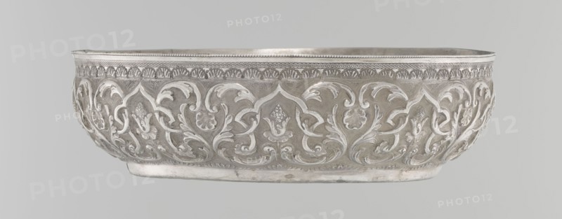 Bowl, purchased provenance noted, Islamic art, Turkish, Ottoman dynasty 1299–1922, geometric motifs and plant ornament, tugra of Abdulhamid II Ottoman sultan; 1842–1918, analogy noted, dated 1876–1909, Turkey, vessel, height 5.6 cm, diameter 19.0 cm, A shallow, oval metal bowl with an ornate raised floral and foliate pattern wrapping around its exterior, showing curved leaf motifs and stylized blossoms in relief; the rim features a patterned border with a small beaded edge and repeated decorative elements beneath it; the bowl rests on a slightly raised base and has a smooth interior visible at the top; the visible colors are shades of silver and gray with subtle variations in tone.