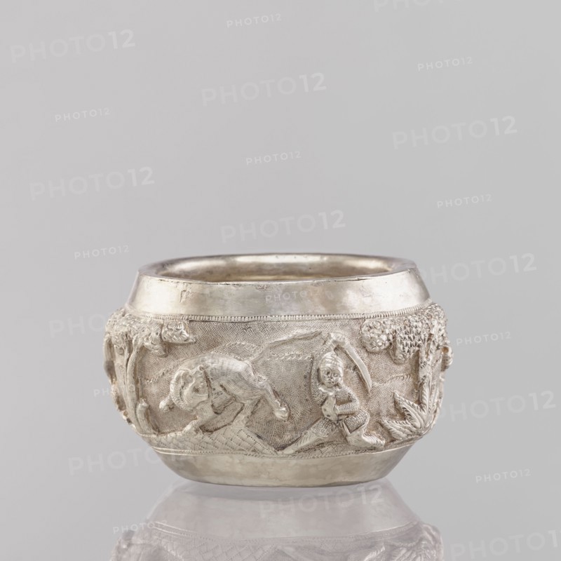 Bowl, South Asian, Indian, applied art, 19th/20th century, India; vessel depicting hunting iconography: tigers, horses, elephants, galloping rider on horseback, warriors, plants; weight 155 g, height 5.4 cm, diameter 8.0 cm., A small round vessel with a metallic silver surface sits against a pale gray background, showing a band of raised sculpted scenes around its midsection with a textured backdrop; the relief includes a figure in motion near a reclining animal, tree-like forms and foliage, and a smooth polished rim and base that reflect light, with overall colors of silver, light gray, and subtle darker gray shadows.