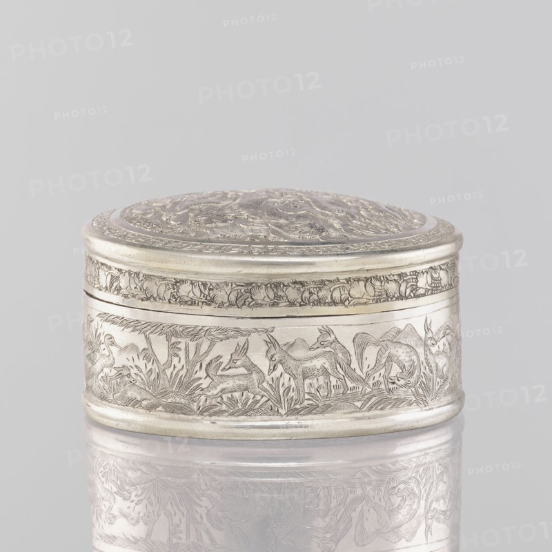 Box with lid; creator, 19th century India; Indian art; materials: tin/container vessel; dimensions: height 3.5 cm, diameter 6.5 cm, weight 105 g. Decoration: animals—lions, antelopes, jackals, cranes—trees, plant and geometric ornaments; applied decoration. Function: box/vessel., The image shows a small oval metal box with an engraved lid and sides, the metal appearing silver and pale gray with darker etched lines; the lid has ornate raised and incised decorative patterns, the rim and banding around the top and bottom are smooth and rounded, and the side surface features an engraved scene of animal figures and vegetation rendered in fine line work; the box rests on a reflective pale surface that mirrors its underside, and the background is a uniform light gray.