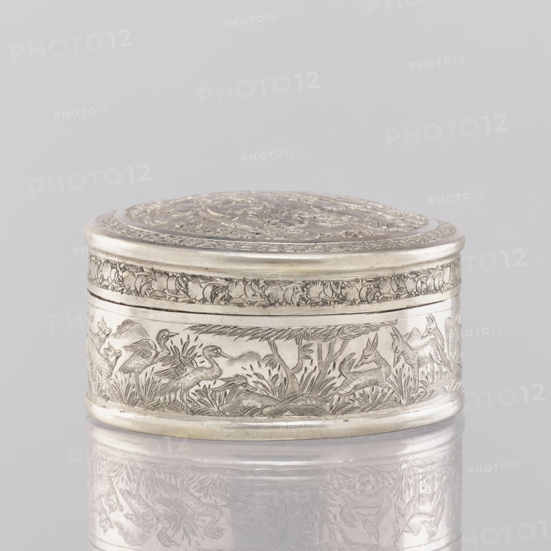 Box with a lid, 19th century, India. Painted metal vessel height 3.5 cm, diameter 6.5 cm, weight 105 g decorated with animals—lions, antelopes, jackals, cranes—trees, plant and geometric ornaments., A small round metal box with a decorated lid sits against a pale gray background, the box and its reflection showing a silver color with engraved scenes around the sides and top; the engravings depict animals and foliage in a darker etched silver tone that contrasts with the smoother silver surface, and a band of floral or leafy ornament wraps near the top edge, while the lid features concentric decorative designs in similar silver hues.
