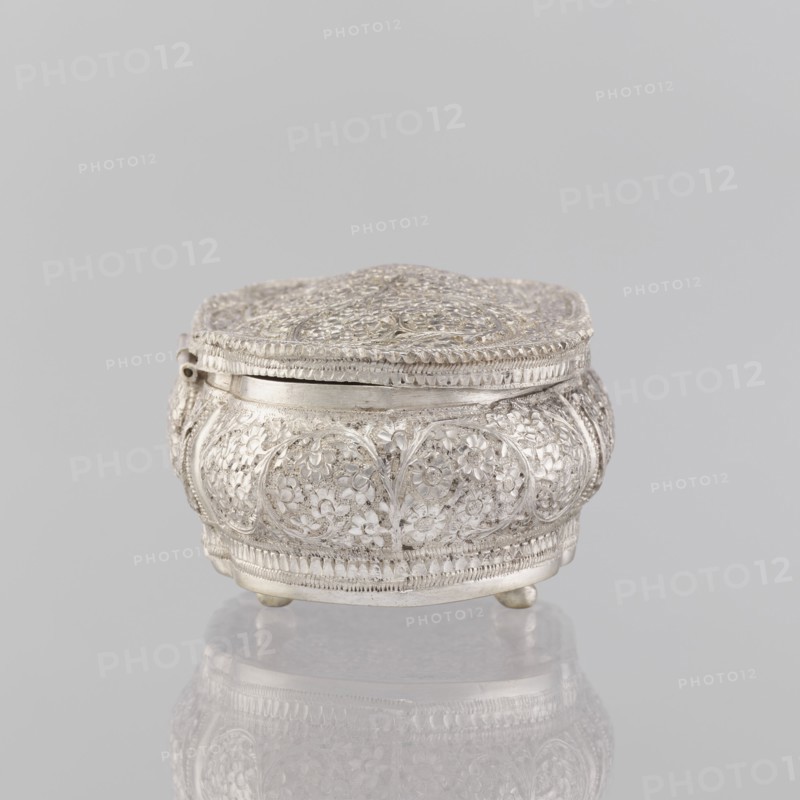 Box with lid, Indian, 19th/20th century, India; plant ornament; height 4.5 cm, width 12.5 cm, depth 6.5 cm, weight 151 g., A small ornate metal box with a domed lid sits on a reflective surface against a plain light gray background; the box is silver in color with intricate raised floral and vine patterns covering its rounded sides and top, a delicate braided edge around the lid, a hinge on one side, and short rounded feet lifting it slightly above the surface, with the silver tones reflected faintly beneath it.