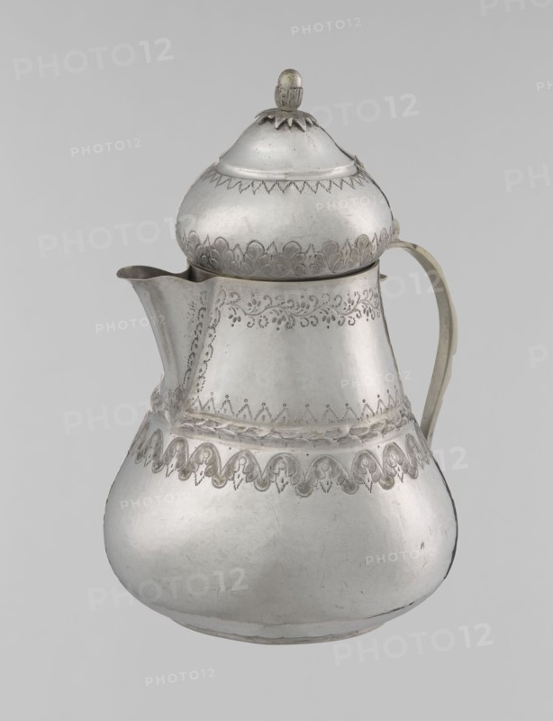 Teapot, 19th century, Turkey; Islamic art, Turkish, Ottoman dynasty 1299–1922; teapot vessel, weight 320 g, height 17.0 cm, diameter 10.9 cm; materials/technique not specified; decoration: geometric motifs, plant ornament, zigzag, lambrequin., A silver-colored metal pitcher with a rounded base, a narrower midsection, and a domed lid topped by a small finial, showing engraved decorative patterns around the upper body, midsection, and near the base; it has a curved spout on one side and a curved handle on the opposite side, with the metal surface showing slight tarnish and subtle scratches, and the overall background appearing light gray.
