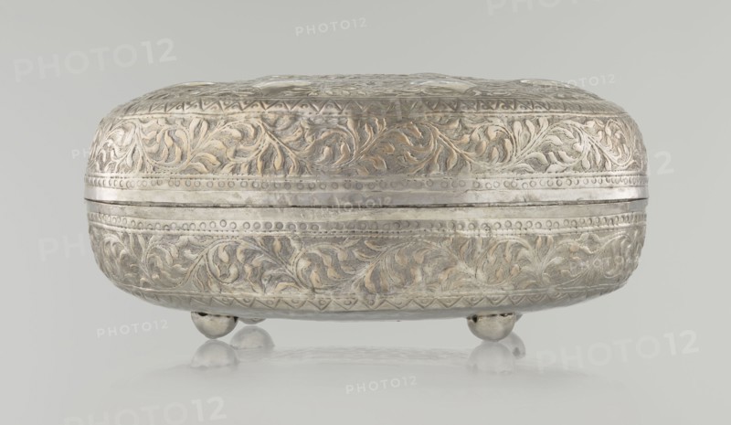 Box with lid; maker, Islamic, Turkish, Ottoman dynasty 1299–1922; 19th century; plant and geometric motifs; materials/technique: varia; dimensions: height 9.0 cm, width 19.5 cm, depth 15.7 cm; weight 210 g., A rounded, ornate metal box with a hinged lid rests on a group of small rounded feet; the surface shows detailed raised floral and vine patterns with textured backgrounds and banded decorative borders. The object appears in silvery and pale gold tones with areas of darker gray patina, set against a light gray background and reflected faintly on a smooth surface beneath it.