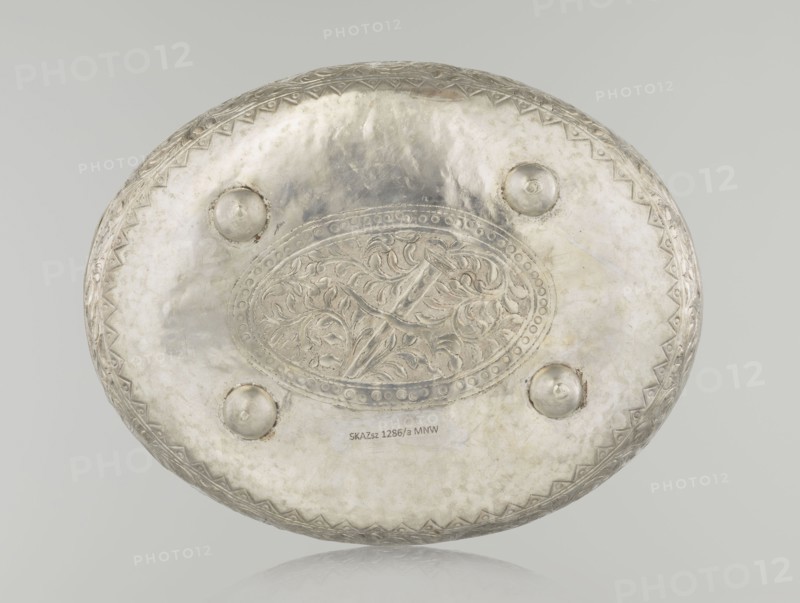 Box with lid, Islamic art, Turkish, Ottoman dynasty 1299–1922, plant motifs, geometric motifs, 19th century, Turkey, varia; height 9.0 cm, width 19.5 cm, depth 15.7 cm, weight 210 g, An oval metal object with a reflective silver surface fills the image, showing an embossed central oval panel with intricate swirling leaf-like patterns and a raised decorative border, surrounded by a smooth area punctuated by a few rounded protrusions; the edge has a repeating triangular zigzag motif, and the overall colors visible are silver, pale gray, and hints of darker gray patina.