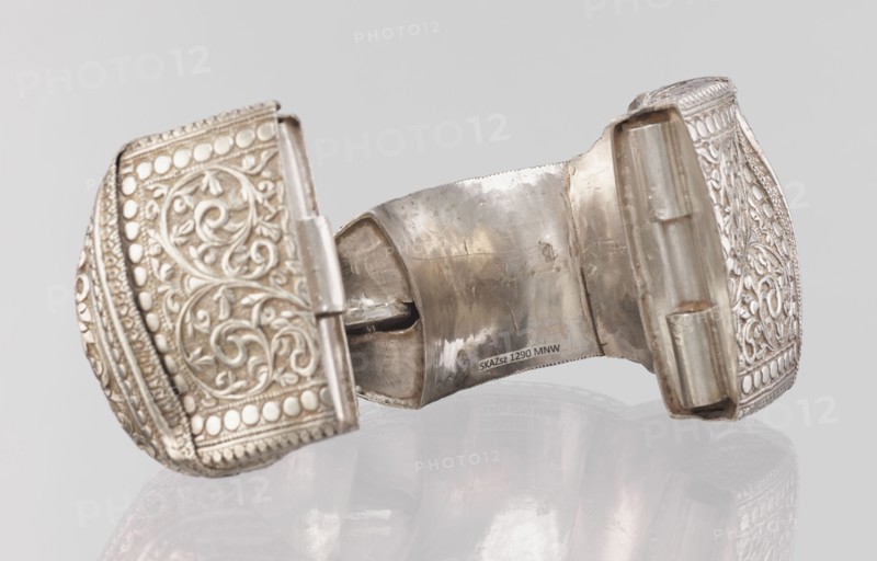 Bracelet, South Asian Indian 19th–20th century. Ring-shaped bracelet of metal jewelry, decorated with a stylized tendril within a rectangular field framed by pearls and tendril motif; pin closure. Dimensions: height 11.4 cm, width 8.5 cm, diameter 12.5 cm; weight 460 g., plant ornament, geometric ornaments., A close-up view of a metallic hinged object with ornate embossed patterns on its two outer panels, showing swirling floral motifs and rows of raised round dots, with a worn central interior surface that is reflective and slightly tarnished; colors visible include silver, gray, off-white, and subtle brownish tones on the metal, set against a pale gray background with a faint reflection of the object below.