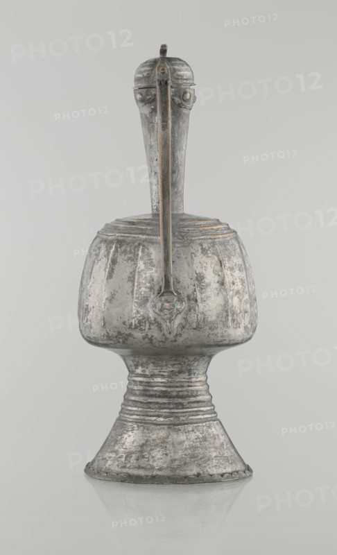 Jug aftabe, 19th century North Caucasus; Islamic art; aftabe water jug; vessel; height 30.0 cm, length 14.8 cm, diameter 12.0 cm, A single metallic vessel with a tall narrow neck rising from a rounded body and flared base is centered against a light gray background; the surface of the object shows a worn, textured finish in shades of silver and gray with darker patches and subtle hints of brownish tarnish; a straight, vertical handle runs from the top of the neck down the front of the body and appears slightly darker with thin lines of brown accent; the rounded body has vertical fluted ridges and a small decorative shield-shaped element where the handle meets the body; the flared pedestal base features concentric horizontal bands and a scalloped edge, and the entire piece is reflected faintly on the smooth surface beneath it.