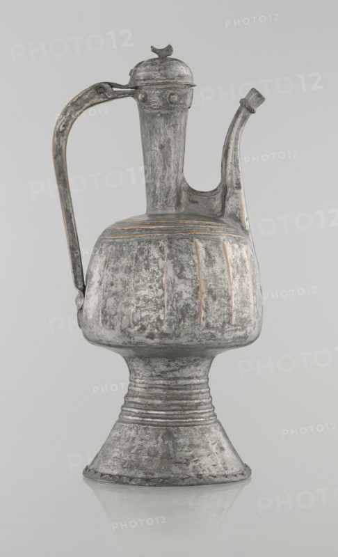 Jug aftabe, unknown provenance; Islamic art; 19th century; North Caucasus; jug/aftabe for water; vessel; height 30.0 cm, length 14.8 cm, diameter 12.0 cm, A metal pitcher with a tall narrow neck, a curved spout, and a high arched handle resting against the side, standing on a flared pedestal base; the surface shows worn, textured patina with areas of roughness and faint vertical ridges around the bulbous body, and a domed hinged lid topped by a small decorative finial; colors visible include shades of gray, silver, and hints of rusty brown.