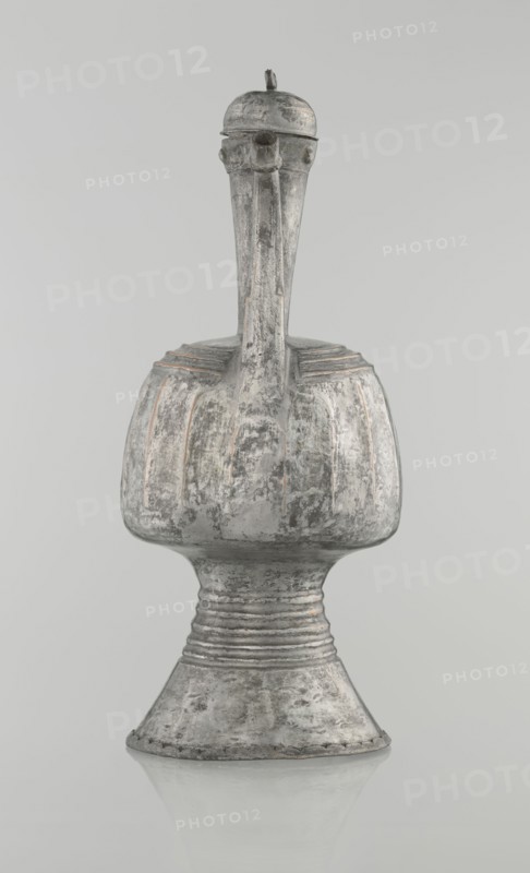Jug aftabe, unknown provenance; Islamic art; 19th century; North Caucasus; jug aftabe for water; vessel; height 30.0 cm, length 14.8 cm, diameter 12.0 cm., A single weathered metal vessel with a tall narrow neck and a bulbous body set on a flared pedestal base, showing a rough, pitted surface texture and areas of worn finish; the form has subtle linear ridges around the shoulder and neck, a rounded lid with a small protrusion on top, and small rounded projections near the lid, all rendered in shades of gray with hints of muted brown and pale metallic highlights against a pale gray background.